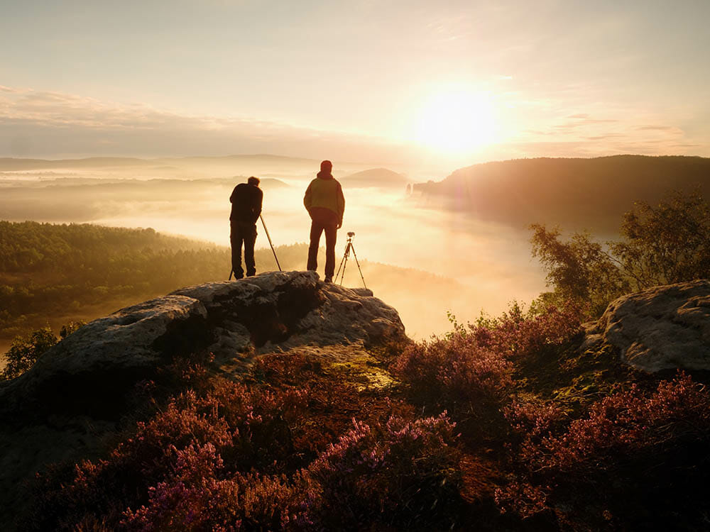 Photographers stay on cliff and takes photos. Dreamy foggy mountains, orange misty sunrise in a beautiful valley below