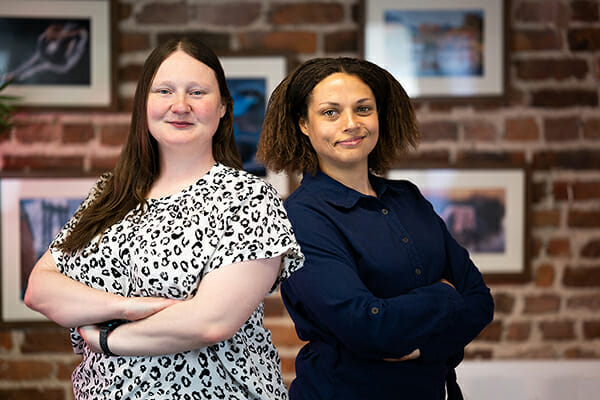 Two ladies stood back to back against a brick wall with pictures on