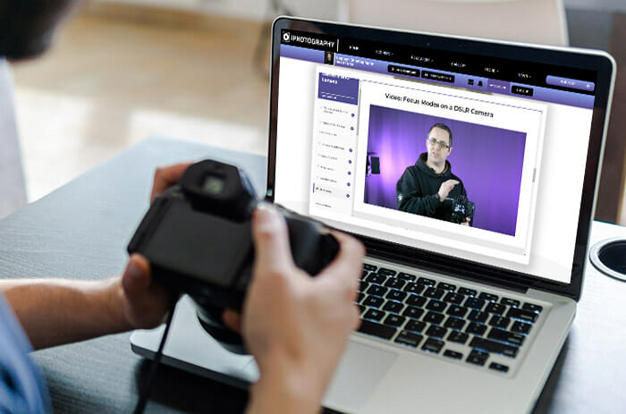 Over the shoulder point of view of a person holding a camera while looking at a laptop which has a photography course on screen
