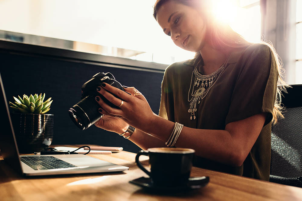 Young woman looking at camera while working on laptop. Photographer with her camera and laptop on her desk.
