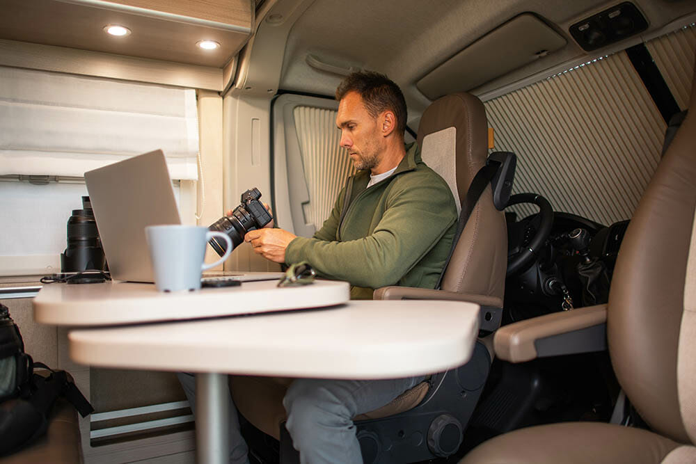Caucasian Man in His 40s Sitting in a Modern Camper Van In Front of a Laptop with a Camera in His Hands, Transferring Recently Taken Landscape Photos to His Computer. Travel Theme.