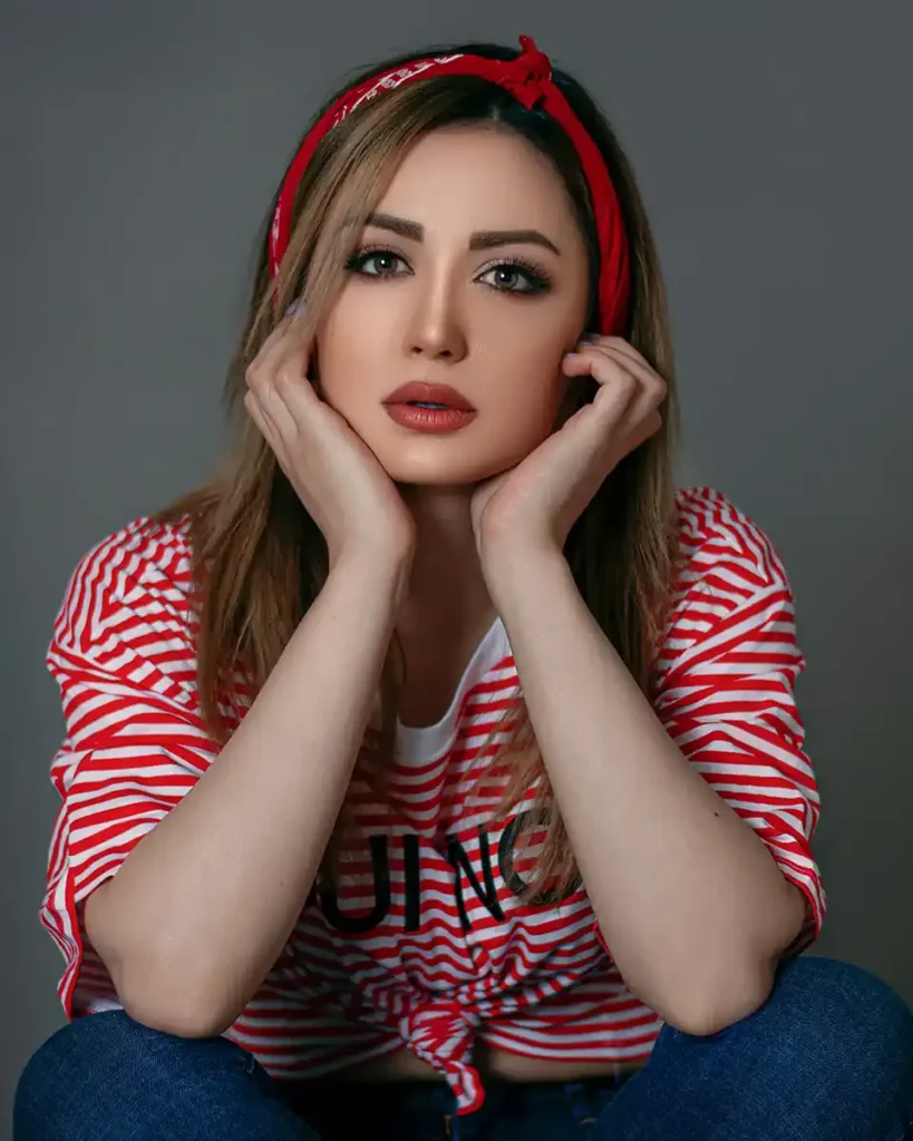 A young lady in a red and white striped top looking at the camera with hands on her face wearing a red hairband