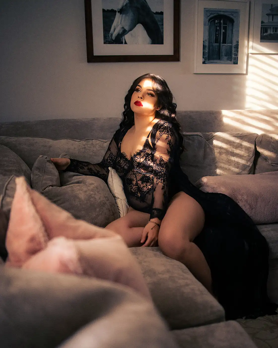 A woman in a black lace dress looking out a window sat on a sofa