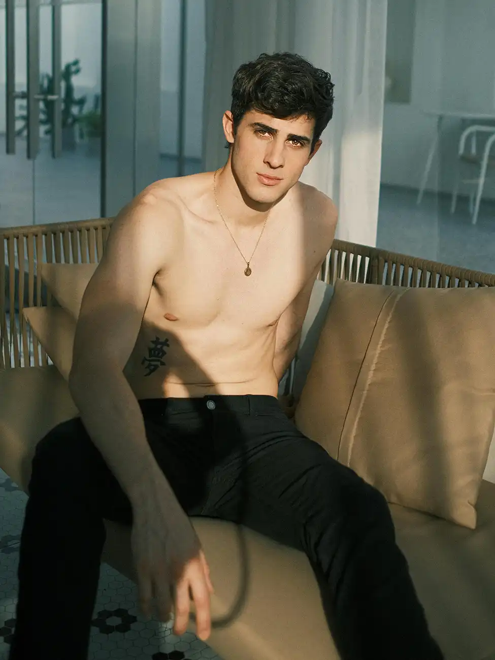 a topless white man in black trousers sat on a beige couch