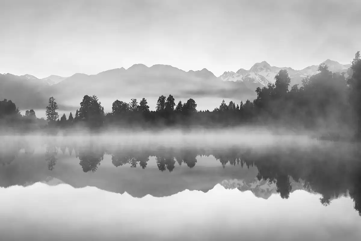 Black and white misty landscape over a lake with trees and mountains in the background