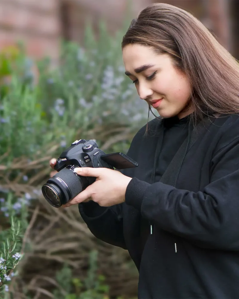 a young female photographer in a black jumper holding a camera taking a photo of plants