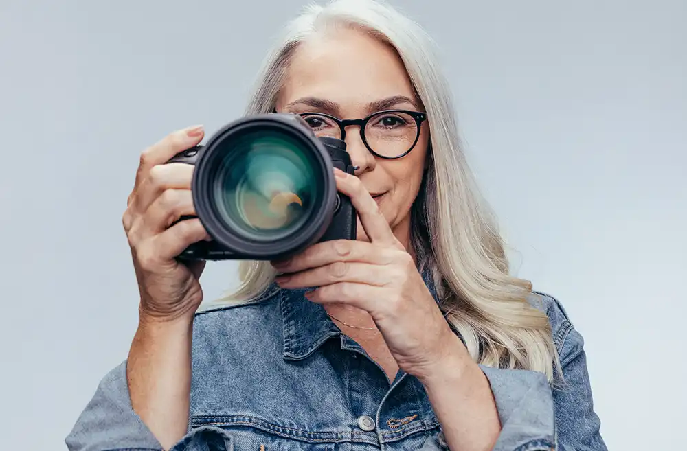 a lady, wearing glasses, with white hair holding a camera wearing a denim jacket