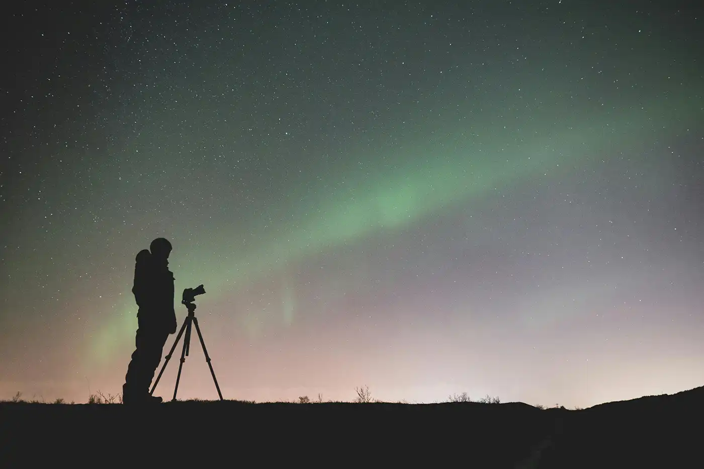 a man standing in silhouette in front of a camera on a tripod taking photos of the northern lights