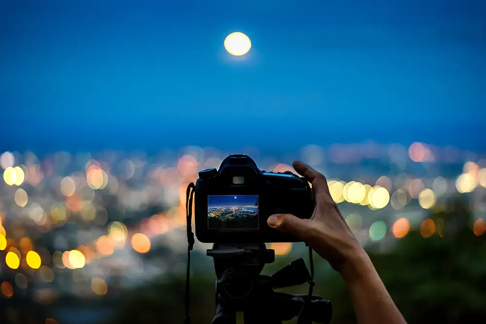 A camera up on a hill at night taking a photo of a blurry moon in the distant background
