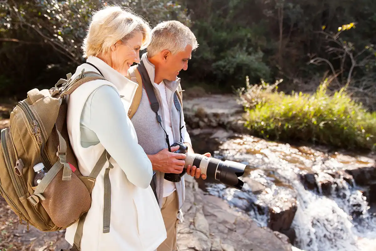 A male and female photographer hold a camera outdoors looking down at a waterfall