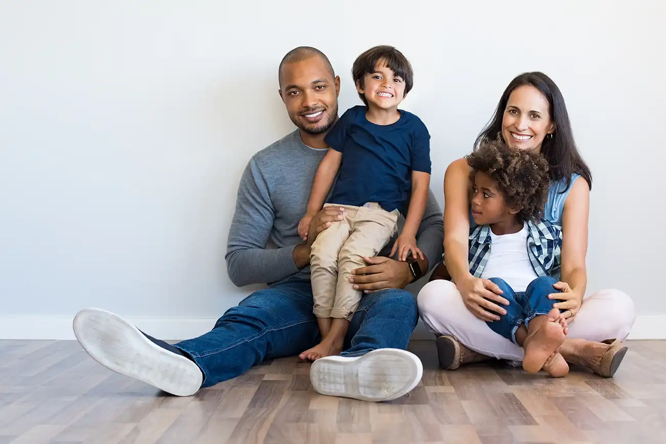 A family sat on a wooden floor against a white wall with two children on their laps